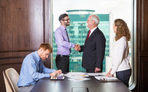 Smiling senior and young businessmen standing at head of table and shaking hands. Businesswoman is standing at table and looking at them. Another businessman is sitting at table and making notes.